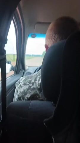The video shows the interior of a car, from the perspective of a passenger in the back seat. The driver, a man with light-colored hair, is wearing a camouflage patterned shirt and is seen from behind. He is focused on the road ahead, which appears to be a dirt road stretching into the distance. The car is traveling through a rural landscape with fields on either side. The weather appears to be overcast, with a bright sky visible through the windshield. The overall scene suggests a journey through the countryside in Podlubovo, Russia. There is no visible text in the video.