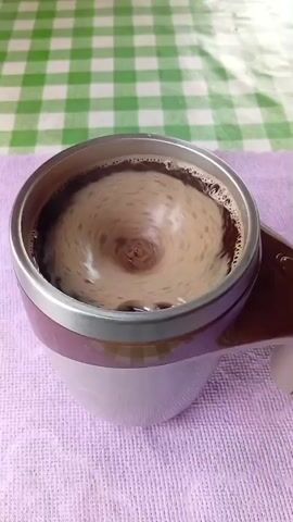 A self-stirring mug is shown from a top-down angle. Inside the mug, a brown liquid is rapidly swirling, creating a vortex in the center. The mug itself is silver with a brown rim and handle. The mug is sitting on a lavender-colored cloth, which is on top of a table covered with a green and white checkered tablecloth. A hand is visible on the right side of the frame, holding the mug by its handle. The video appears to be a close-up shot of the self-stirring mechanism in action.