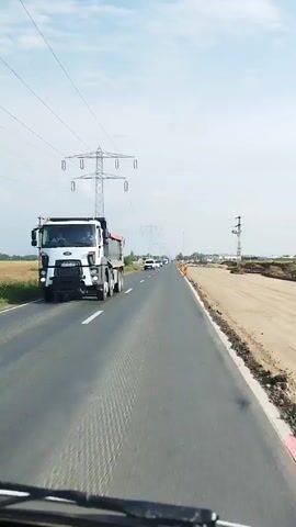 The video shows a view from a vehicle driving on a paved road. There are multiple lanes on the road, with traffic moving in the distance. On the right side of the road, there is a construction site with dirt and debris. Power lines and towers are visible in the background. The sky is cloudy.