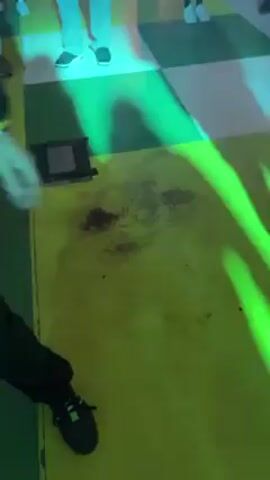 The video shows a person wearing a black jacket, yellow hoodie, blue cap, and reflective yellow glasses holding a cup with a Halloween theme. They are standing on a yellow and green checkered floor, smiling and pointing. The floor has a dark reddish-brown stain on it. The person is surrounded by other people in what appears to be an indoor venue with multiple levels. The lighting is predominantly green and blue, and there is a hint of fog or haze in the air. The music is loud and electronic.