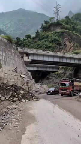 The video shows a damaged concrete retaining wall on the left side of a dirt road. The wall has large sections that have collapsed, with debris scattered at its base and on the road. In the background, there is a multi-lane bridge spanning over the road. Several vehicles are visible on the road, including a red truck, a maroon car, and another car. The road appears to be in a mountainous region with lush green vegetation on the slopes of the mountains. The sky is overcast, suggesting cloudy weather. The overall scene depicts a challenging infrastructure situation in a natural environment.