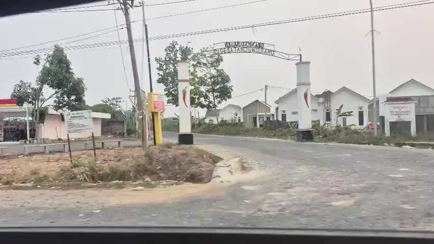 The video shows a street view from a moving vehicle. On the left, a red and yellow minimarket with cars and motorcycles parked in front is visible. Further down the street, a small shack with a sign that reads "SHOES VAGANZA" is present. On the right side of the street, there are houses and an archway. The sky is overcast.