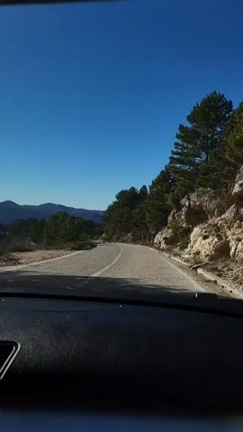 From inside a vehicle, the camera is pointed forward out the windshield as the vehicle drives on a narrow road with a center line. The sky is a clear blue. Evergreen trees grow thickly on the right side of the road, next to a rocky embankment. On the left side of the road, evergreen trees grow more sparsely. In the distance are mountains. A yellow road sign is visible on the right side of the road. The dashboard of the vehicle is black. A man is speaking in Russian.