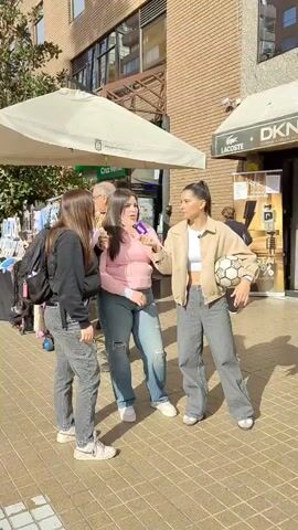 Three young women stand on a paved outdoor area, possibly a sidewalk or plaza, in front of a brick building with storefronts. The woman on the left wears a black hoodie and gray jeans, carrying a black backpack. The woman in the middle wears a pink long-sleeved top and distressed blue jeans, and holds a purple object, possibly a microphone, in front of her face. The woman on the right wears a tan jacket over a white crop top and gray wide-leg jeans, and is holding a soccer ball under her left arm. She is speaking into a microphone. The women appear to be interacting with each other, possibly for a video recording or interview. In the background, a white awning is visible, as well as storefront signs for DKNY and Lacoste. Other people can be seen walking in the background. The ground is paved with square tiles, and sunlight creates shadows on the ground.
