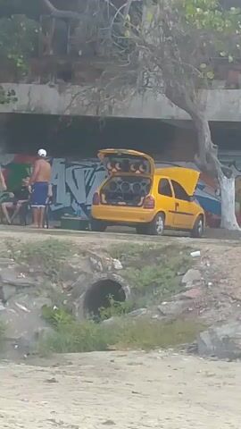 A yellow car with its trunk open and hood slightly ajar is parked. The trunk is filled with speakers. Two people are standing near the car, and another person is sitting. The car is parked in front of a graffiti-covered wall. There is a tree growing in front of the wall and the car. The area appears to be outdoors and possibly near a beach, as indicated by the sandy ground in the foreground and vegetation in the background. There is also a concrete pipe visible in the foreground.