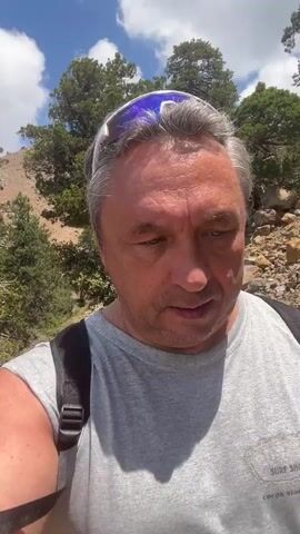 A man with grey hair, wearing a grey t-shirt, sunglasses, and a backpack, is standing outdoors on a dirt path. He is looking directly at the camera and appears to be speaking. The background shows a rocky, mountainous landscape with scattered evergreen trees and a blue sky with white clouds. The man's left arm is visible, and the strap of his backpack is on his shoulder. The overall impression is of a person on a hike in a natural, elevated environment.