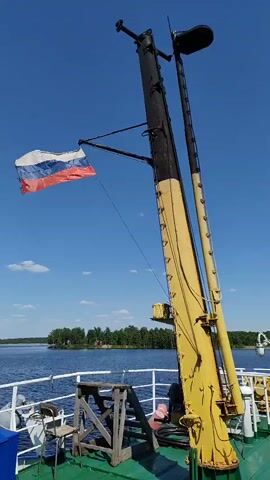 The video shows a boat on a lake with the Russian flag flapping in the wind. The foreground features a pole, painted yellow and black, with a light on top. A chair sits nearby. The background reveals a clear blue sky and a tree-lined shore across the water.
