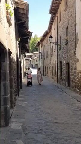 A man and a woman are walking down a cobblestone street in a village. The man is pushing a stroller with a baby inside. The woman is walking beside him, carrying a handbag. The buildings on either side of the street are made of stone and have a rustic appearance. The sky is clear and blue.