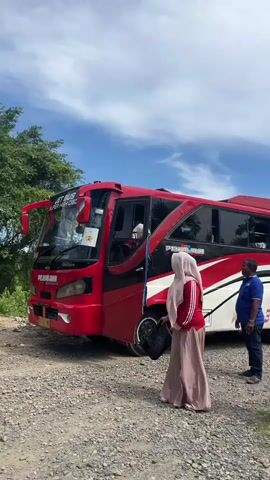 A group of people, mostly women wearing hijabs, are gathered near a red bus. The bus appears to be parked on a gravel road next to a paved path. In the background, a large building with a tall minaret, resembling a mosque, is visible. The sky is blue with some clouds. Some individuals are standing near the bus, while others are walking away from it. One person is holding a white object, possibly a phone or a cup. The environment suggests an outdoor setting, possibly a tourist destination or a place of worship.