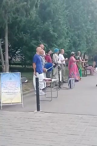 A group of people are standing in a line next to a sidewalk with a paved surface and a grassy area with trees in the background. Some of the people are holding musical instruments, and one person is playing a drum. A sign with text and a symbol is standing to the left of the group. Another person is walking towards the right side of the frame, away from the group. The lighting suggests it is daytime.