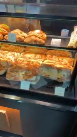 The video shows a bakery-cafe at an airport.  The display case is filled with pastries and sandwiches, including croissants, various types of flatbreads, and sandwiches on baguette bread. Behind the display case, employees wearing white chef hats prepare orders. A line of customers is waiting at the counter.  The cafe is labeled "PAUL."  The floor has a black and white checkered pattern. Large windows offer a view of the airport terminal.  People are seated at tables outside the cafe.