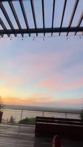 A video shows a wooden deck with benches and railings overlooking a body of water under a sky with clouds tinged pink and orange from a sunset. String lights are visible overhead. The deck appears wet, possibly from rain, with water puddles reflecting the sky. The video pans from the sky and water to the deck and then shows a person's legs and feet wearing burgundy sneakers and red socks, suggesting they are sitting or lying down. The camera then pans back towards the water and sky.