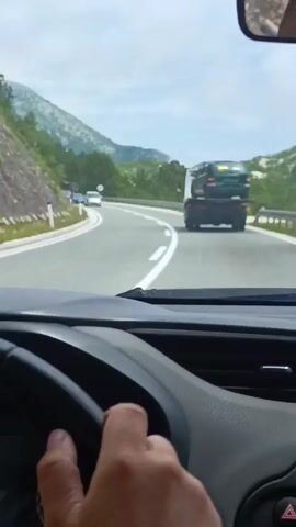 The video is taken from the perspective of a person driving a car. The driver's hands are visible on the steering wheel. The road is a two-lane highway curving through a mountainous, forested area. Other vehicles are visible on the road, including a tow truck carrying a car, a black SUV, and other cars in the distance. The sky is cloudy.