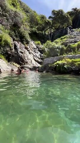 A group of people swim and interact in a clear, green body of water. The water appears to be a natural pool or river with rocks visible on the bottom. The surroundings are lush and green, with rocky slopes and trees rising up from the water's edge. Sunlight streams down, creating dappled light on the water's surface. The people are of various ages and genders, and they are engaged in activities like swimming, talking, and playing. In the background, a waterfall can be seen cascading down the rocks. The overall atmosphere is one of leisure and enjoyment in a beautiful natural setting.