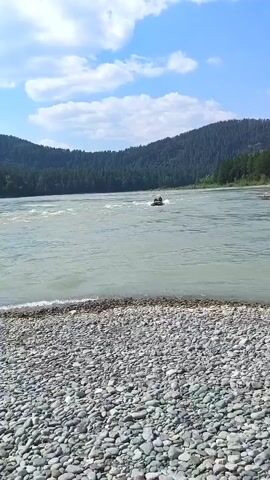 A small inflatable boat with several occupants navigates a wide, fast-moving river. The boat is in the middle of the river, leaving a white wake as it moves. The river is surrounded by densely forested mountains. In the foreground, a rocky shoreline is visible. The sky is blue with scattered white clouds.