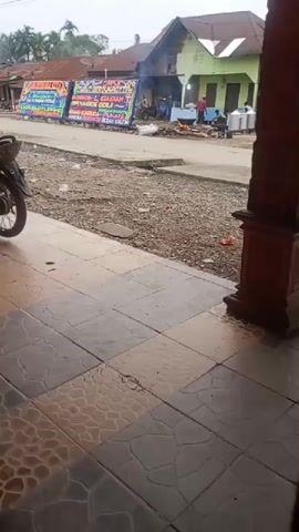 The video shows a street scene, possibly in Southeast Asia, with a few buildings in the background and a sidewalk in the foreground. There are several large floral arrangements displayed on the street, which appear to be for a funeral or a similar event. A few people are gathered around, and there is a small fire burning in the middle of the street. A motorcycle is parked on the left side of the frame. The overall impression is one of a community gathering for a solemn occasion.