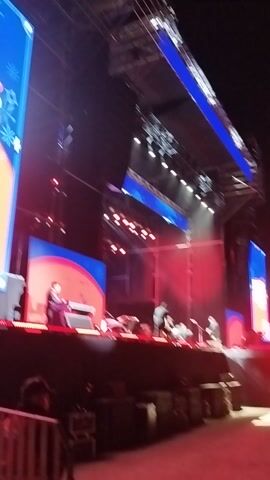 The video captures a lively stage performance at night, likely an outdoor concert or large event, from a high vantage point looking down. The setting features a prominent stage illuminated by an array of bright red and blue stage lights.

On the stage, several individuals are visible, appearing to be musicians or performers. Some are standing, while others are seated. One person is seated in front of a console or keyboard, actively engaging with a large screen behind them. Other performers are visible further back on the stage, suggesting a band or ensemble setup. Equipment like drum kits and other musical instruments are discernable in the background.

Large LED screens are a central feature of the stage design, displaying dynamic and colorful graphics. One screen prominently features a red circular shape against a blue background, serving as a backdrop for the seated performer. Another screen, visible in the bottom left, displays blue and orange stylized graphics, with the word "Grace" clearly legible among other decorative text. The overall atmosphere is vibrant and energetic, characterized by the pulsating lights and the evident live performance. The video's perspective suggests it was filmed from an elevated position within the audience at a venue in Maipú, Chile.
