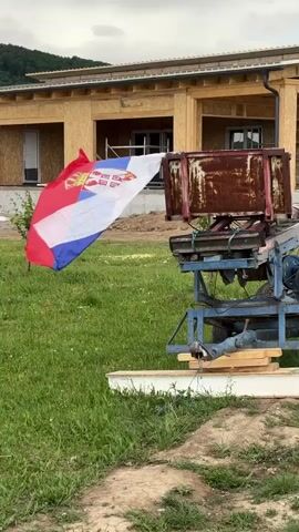 The video shows a flag of Serbia waving in the wind in front of a wooden building under construction. The building is framed but not yet finished. There is scaffolding visible through the window openings. In the foreground, there is some kind of machinery, possibly for construction, with a rusty container attached. The ground is covered in green grass. The sky is overcast.