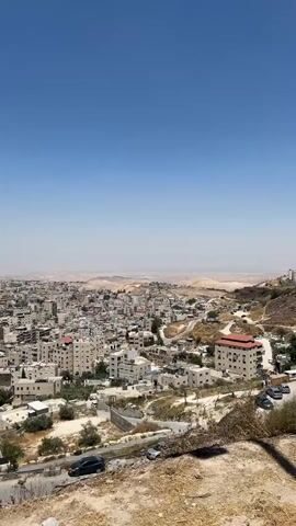 The video pans across a view of a dense urban area nestled within a hilly, arid landscape under a clear blue sky. The buildings are predominantly made of light-colored stone or concrete, with many featuring flat roofs and balconies. The architecture suggests a Middle Eastern or Mediterranean setting. Streets wind through the town, with a few cars visible, including dark-colored sedans and a white vehicle. The vegetation is sparse, consisting of scrubby bushes and some trees. In the distance, rolling hills and a hazy horizon suggest a vast, dry expanse. The overall impression is of a sun-drenched town built on a challenging, rocky terrain.