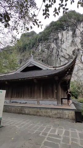 The video shows a wooden building with a curved roof, likely a temple or traditional structure, set against a backdrop of a large, rocky cliff covered with some vegetation. Two people, a man and a woman, are seen walking out of the building. The man bends over, possibly adjusting his shoelaces or picking something up. An older woman in a red jacket walks past the camera. The area around the building is paved with stone tiles. There are trees, some with sparse leaves, scattered around the area. The sky is overcast. A sign and a trash bin are also visible in the scene.