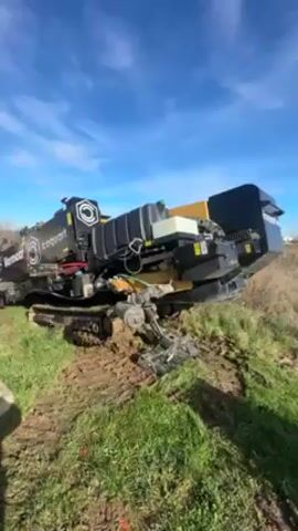The video shows a large black and yellow Vermeer Coquart horizontal directional drilling machine on a grassy verge next to an asphalt road. The sky is blue with scattered clouds.  

A white truck with a red frame carrying a load of black pipes is parked on the road next to the drilling machine. Two people wearing orange vests are standing near the back of the truck. A tire is visible on the ground next to the truck. Traffic is visible in the distance on the road.