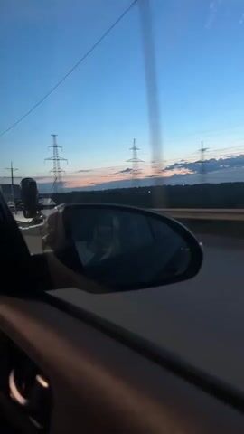 The video shows a view from inside a moving vehicle looking out the passenger side window and in the rearview mirror. The time of day is dusk or dawn, with a blue sky and soft pink and orange hues on the horizon. Tall electrical towers and power lines are visible in the distance. In the rearview mirror, a person is reflected, holding a mobile phone to take a selfie. The vehicle is on a road, likely a highway, with a guardrail visible. The camera is shaky, indicating movement.