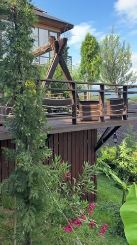 This video shows a scenic view of a wooden balcony with chairs and a rustic wooden structure. The balcony is attached to a house that appears to be built into a hillside. In the foreground, there is lush green foliage and pink flowers. The background reveals a charming village with A-frame houses surrounded by trees and rolling hills. The sky is blue with scattered clouds, suggesting a pleasant day. The camera pans slightly, offering a wider perspective of the tranquil landscape.