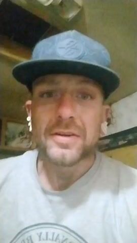 Here's a concise description of the video:

The video features a close-up shot of a man with fair skin, a short beard, and light-colored eyes. He is wearing a gray t-shirt and a blue baseball cap with a dark brim and a white circular logo on the front. He has a nose ring and stretched ear piercings with dangling earrings. The background is somewhat blurry and includes what appears to be a wooden cabinet. The man is speaking directly into the camera.