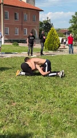Two men are grappling on a grassy lawn. The man on top is shirtless, wearing black sweatpants and sneakers. The man underneath is wearing a black long sleeve shirt, black sweatpants, and tan sneakers. Both men are on their knees and the man on top has the other in a headlock. There are other people standing in the background watching. In the far background is a two-story brick building with a tree in front of it. The day is sunny and bright.