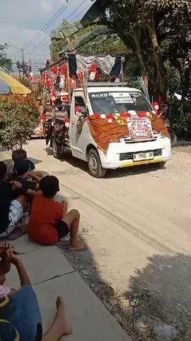 The video shows a white pickup truck decorated with banners, red and white streamers, and fabric draped around the sides, as it drives down a residential street. The truck appears to be part of a parade or procession. Spectators are sitting on the curb watching the truck pass. The environment is a sunny outdoor setting with trees and houses visible.