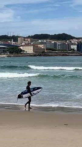A person in a black full-body wetsuit walks along a sandy beach, carrying a surfboard under their right arm. The surfboard is white with a distinctive black and dark blue abstract pattern, possibly resembling flames or tribal designs. A black leash trails from the board. The individual is moving from the right side of the frame towards the left, approaching the water's edge.

The ocean in the mid-ground features moderate, breaking waves with a greenish-blue color. A few other dark figures, likely surfers, are visible further out in the water, beyond the immediate surf zone.

The background showcases a city skyline, characteristic of A Coruña, Spain. Numerous buildings of varying heights and architectural styles, predominantly in light and dark grey tones, line the coast. Behind the urban development, a green, tree-covered hill rises. To the far left of the city view, an elevated structure, possibly a bridge or viaduct, is visible.

The scene is set during a bright day with a partly cloudy sky. The overall impression is one of calm preparation for water sports, with the urban landscape providing a vibrant backdrop to the natural beach environment. No discernible text is visible on the surfboard or elsewhere in the image.