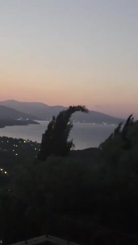 The video displays a panoramic view of a coastal landscape at dusk. The sky is a gradient of pale lavender and orange, with the fading light casting a soft glow. In the foreground, dark silhouettes of trees and bushes dominate the lower portion of the frame, their shapes indistinct against the dim light. To the left, the roof of a white building with a terracotta tile roof is partially visible. Beyond the trees, a body of water, likely a bay or sea, stretches into the distance, reflecting the twilight sky. Across the water, rolling hills and mountains form a layered backdrop, their peaks and slopes softened by the haze of dusk. Along the coastline and nestled in the valleys between the hills, numerous small lights twinkle, indicating a town or village coming to life with artificial illumination as darkness descends. The overall impression is one of a peaceful, serene evening scene, capturing the transition from day to night.