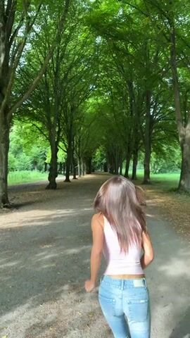 A young woman with long brown hair runs away from the camera down a dirt path lined with tall trees. She is wearing a pink crop top and light blue flared jeans. The trees form an archway over the path, and their leaves are a vibrant green. The ground is covered in fallen leaves. Sunlight filters through the leaves, creating dappled shadows on the path.