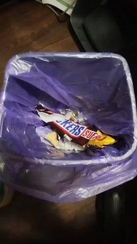 A trash can lined with a purple bag contains a crumpled Snickers wrapper and other debris. The camera slowly moves closer and then farther away from the trash can, providing a detailed view of the contents. The Snickers wrapper is partially torn open, revealing some of the candy bar inside. Other wrappers and pieces of food are also visible in the trash can. The background of the scene is blurry, but it appears to be an indoor setting with wooden flooring.
