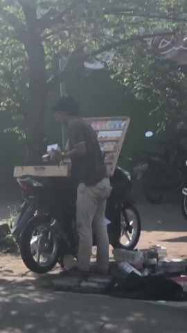 A young man wearing glasses and a dark t-shirt stands next to a motorcycle. He is wearing light-colored pants. The motorcycle has a wooden display rack attached to the back, filled with various items, possibly snacks or small products. The man is reaching into the display rack. The scene appears to be outdoors, with trees and shadows visible. There is also a building in the background, with some text that reads "FREE WI". The ground is paved.