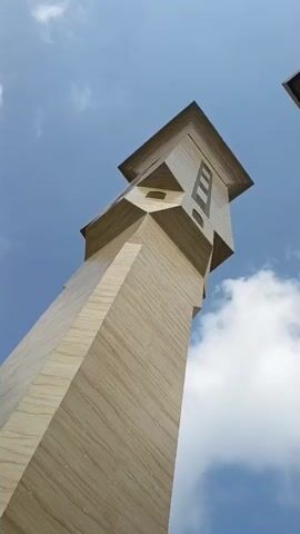 The video shows a low-angle shot of a modern minaret against a bright blue sky with scattered white clouds. The minaret is tall and slender, with a light-colored, textured exterior that appears to be made of stone or a similar material. It has a geometric design with angular protrusions and recessed sections. The camera slowly zooms out, revealing more of the sky and a portion of another building on the right side of the frame. The lighting suggests it is daytime, with the sun high in the sky. There are no people or animals visible in the video.