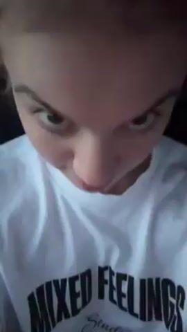 A fair-skinned young adult female is in a vehicle. She wears a white t-shirt with the words "MIXED FEELINGS" and the word "Studio" written below in script. Her hair is light brown and pulled into a high, messy bun. At the beginning of the video, she sticks her tongue out. The video angle is angled from above her. She places her right hand on her cheek and stares at the camera. The interior of the car is visible behind her.