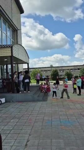 A group of people, including adults and children, are gathered outside a building on a paved courtyard. Some people are standing on steps leading to the building entrance. Several children are seated on the steps. Many of the people are holding colorful balloons. The ground is speckled with colorful powder, likely from a Holi celebration. The sky is blue with fluffy white clouds. The building appears to be a school or public institution. The overall atmosphere seems festive and celebratory.