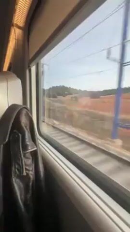 The camera looks out a train window. Outside the window, a landscape of low, scrubby trees and red-tinged soil moves by. Power lines hang above the railroad tracks, visible in the foreground. A leather jacket is hung on the seat next to the window.