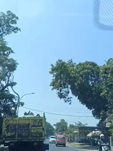 The video shows a view from a vehicle looking forward. The sky is clear and blue. In the foreground on the left is the back of a yellow truck with black graffiti. A red van is visible driving on the road ahead of the truck, followed by a white car. To the right of the road, there are trees, power lines, and some small buildings with awnings. A large tree with green leaves extends over the road from the right side of the frame. The overall impression is a sunny day on a suburban or rural road.