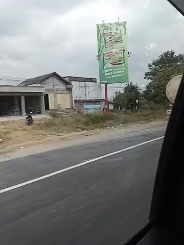 The video shows a view from inside a vehicle traveling along a road. On the left side of the road, there is a billboard with text and images of food products. Adjacent to the billboard are several buildings, some appearing to be shops or houses, with dry vegetation and sparse trees. A large white tanker truck is visible on the road. Further down the road, a smaller white box truck with green lettering is seen. In the distance, other vehicles and trees are present. The sky is overcast. The road is paved with white lane markings. The video captures the movement of these vehicles and the roadside scenery as the filming vehicle progresses.
