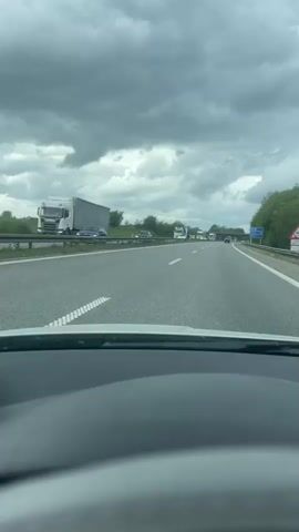 The video shows a first-person perspective from inside a car driving on a highway. The sky is overcast with dark clouds. Several trucks can be seen on the opposite side of the highway, traveling in the same direction. The camera is static within the car. The overall tone is neutral, documenting a typical highway drive on a cloudy day.