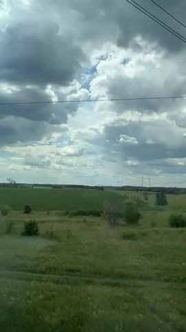 The media file shows a view from a moving vehicle, likely a train, looking out at a rural landscape. The scene is dominated by a vast, green field of crops stretching towards the horizon, under a dramatic sky filled with heavy, grey clouds interspersed with patches of blue sky. Tall, modern electricity pylons with power lines crisscross the upper portion of the frame, indicating infrastructure in the distance.

In the far distance, several plumes of white smoke rise from what appears to be industrial chimneys, suggesting the presence of a factory or power plant. The foreground is filled with wild, green grass and scattered bushes and small trees, some with yellow wildflowers blooming. The overall impression is of an overcast day, possibly late afternoon or early evening given the light. There are no people or discernable objects in the foreground, and no text is visible. The overall mood is serene and vast, with a hint of industrial presence on the horizon. The location context provided is Sof'ino, Russia.