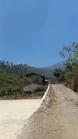 A low-angle shot shows a dirt road leading up a hill. On the left side of the road is a concrete area and a small building with a gray roof. In the background, lush green trees and mountains can be seen under a clear blue sky. The scene appears to be a rural setting on a sunny day.