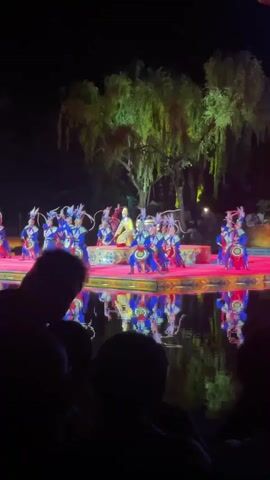 A nighttime performance is in progress on a red stage, featuring around a dozen performers in blue, red, and white costumes and headdresses. The dancers are arranged on a low, ornate platform and are reflected in the water in front of the stage. A tall green tree stands behind the stage. The audience is silhouetted in the foreground.