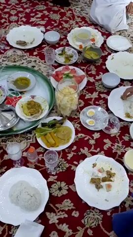 A static, medium shot shows a dining table covered with a red patterned tablecloth. Numerous white plates and bowls are scattered across the table. Many dishes are partially eaten, including rice, chicken, vegetables, and watermelon. There are also small glasses filled with water or other beverages. A couple of trays are placed on the table. The scene implies a meal has taken place.