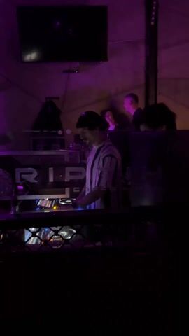 A DJ is working at his station in a nightclub with purple lighting. He is wearing a black beanie and a white and black striped shirt. The booth in front of him has the words "TRIP CAFFE & BAR" written on it. Three people are standing in the background behind the DJ. A large black screen hangs on the wall.