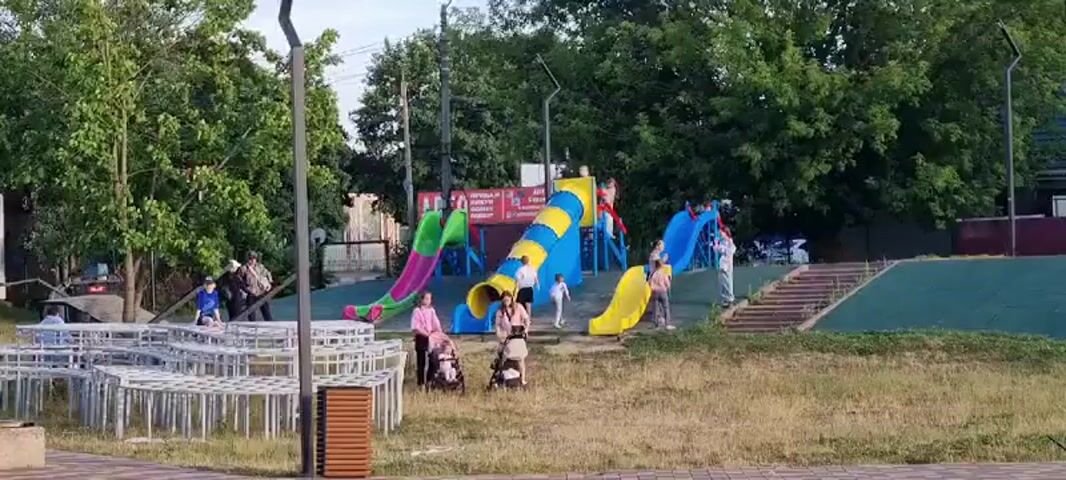 A group of people are gathered at an outdoor playground on a sunny day. The playground features several colorful slides, including a large blue and yellow tube slide, a green and pink slide, and a yellow spiral slide. Children are seen playing on the slides, with some adults supervising. In the foreground, there are white picnic tables and benches, and a brown trash can. The environment appears to be a park or recreational area with trees and grass.