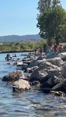 The video shows a scenic view of a body of water with people swimming and wading. The water is clear and blue, with small waves lapping at the rocky shore. In the background, there are lush green hills dotted with buildings and trees. The sky is clear and blue, indicating a sunny day. Several people are visible in the water, some standing, some swimming. On the shore, there are more people walking and standing amongst the rocks. The scene suggests a relaxed, recreational atmosphere at a lake or seaside location.