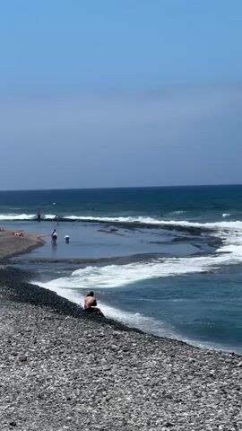 This media file captures a serene day at a coastal location, likely in Arona, Spain. The scene is dominated by the vast, deep blue ocean meeting a rocky shoreline. In the foreground, a pebble beach is visible, with a person sitting on it, facing the water. Further out, the ocean is active with waves breaking, creating a foamy white surf that adds a dynamic element to the tranquil setting.

Beyond the immediate shoreline, the water forms natural pools or inlets, indicated by calmer, shallower areas where the waves recede. Several people are present in these calmer waters, some standing and others appearing to wade or play. In the distance, more figures can be seen near the water's edge and further out in the ocean, suggesting a popular spot for recreation.

The sky above is a clear, bright blue, with a few wispy clouds adding texture to the upper portion of the frame. The lighting suggests it is daytime, likely midday or early afternoon, given the brightness and the shadows. The overall mood is peaceful and relaxing, with people enjoying the natural beauty and the refreshing ocean. There is no visible text in the media file. The location context of Arona, Spain, suggests a Mediterranean or Atlantic coastal environment, fitting with the visual cues of a sunny, seaside atmosphere.